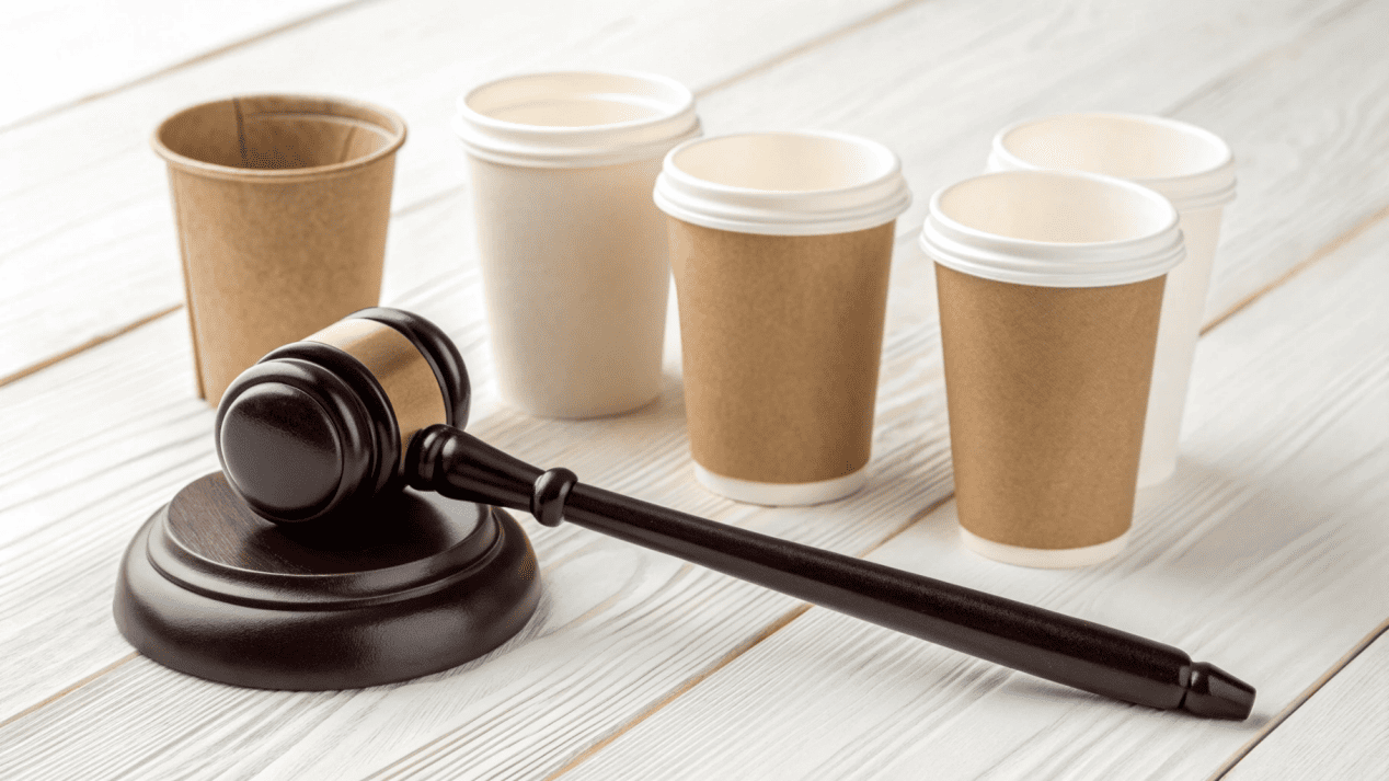 A gavel next to a stack of different sustainable paper cups, symbolizing regulations.