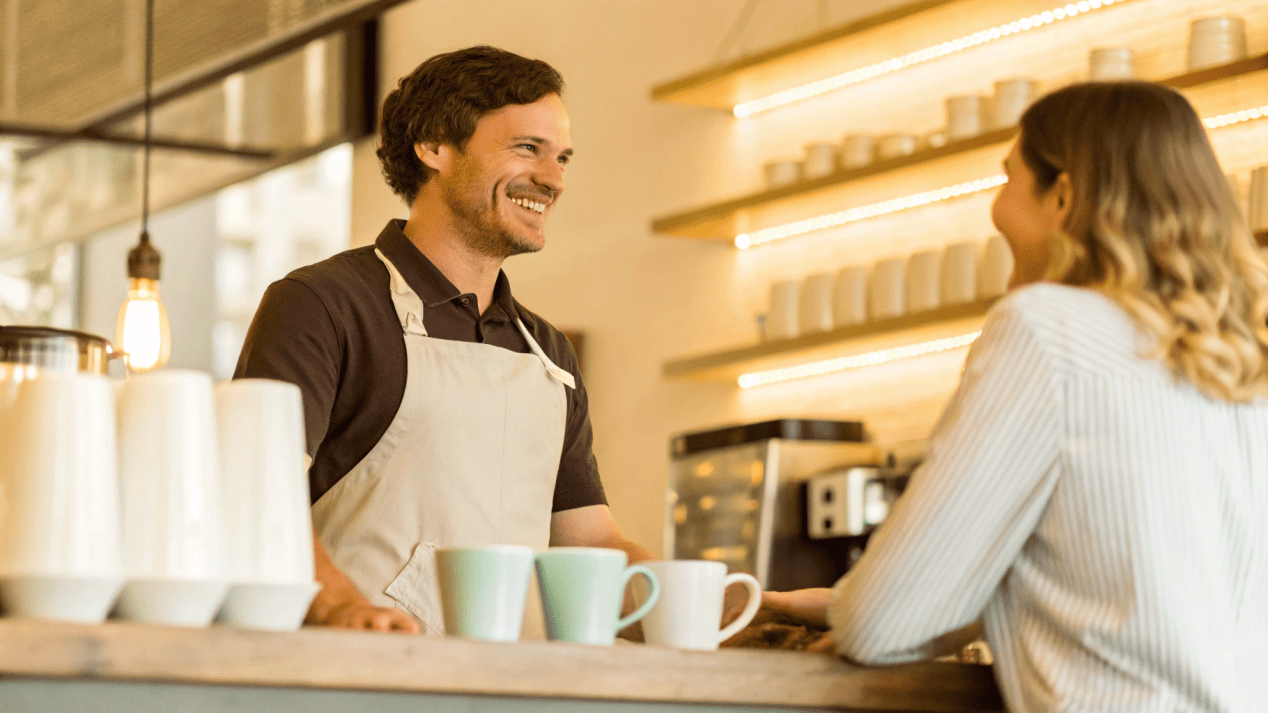 A friendly barista asking a customer, "Is this for here or to go?" with ceramic mugs visible in the background.
