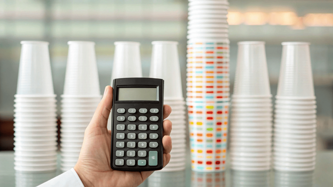 Cost of Plastic vs. Paper Cups A hand holding a calculator with stacks of plastic and paper cups in the background.